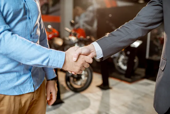 Aperto de mãos entre um profissional de uniforme azul e um executivo de terno, com motos ao fundo